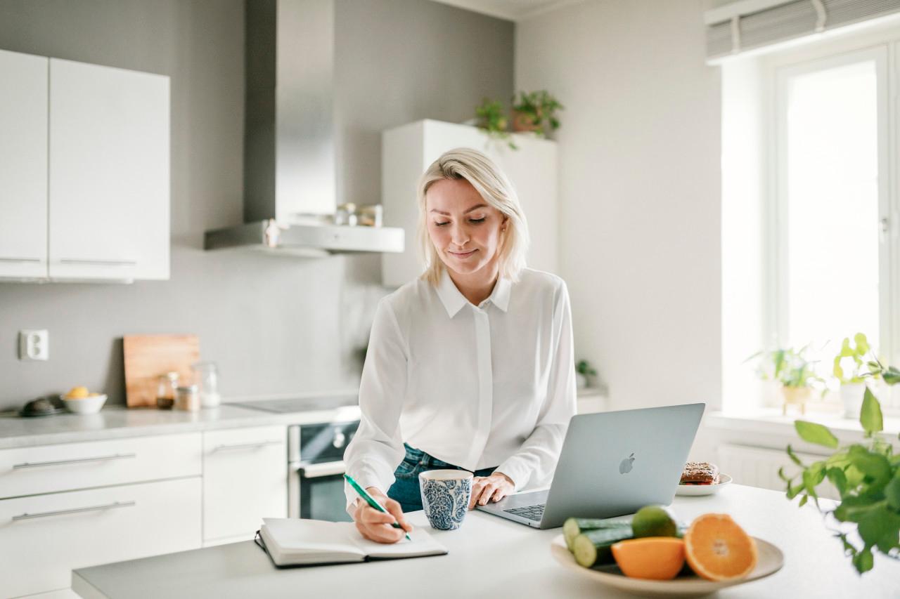 Blond kvinna sitter i ett ljust kök, dricker kaffe, tittar på sin macbook och gör anteckningar